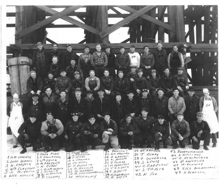Rows of men in work clothes and caps posed before the timber coal trestle, a numbered handwritten name key below